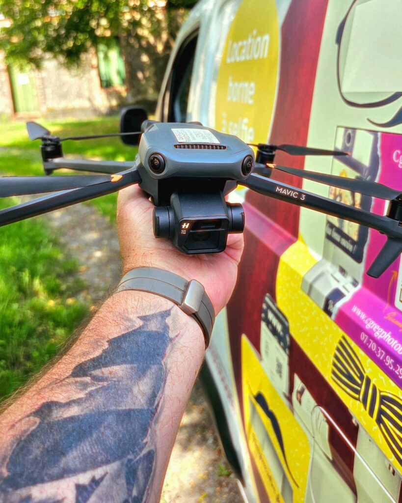 Une main tatouée soulève délicatement un drone DJI Mavic 3. En arrière-plan, légèrement flouté, se devine la silhouette d'un véhicule utilitaire, sur lequel on peut lire une offre de services photo et de location de bornes à selfie.