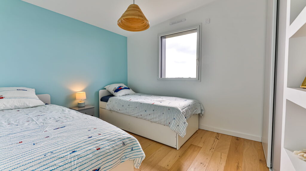 Cette chambre baignée de lumière accueille deux lits simples. Un mur bleu pastel apporte une touche douce et apaisante, tandis que le linge de lit arbore des motifs marins qui rappellent les vacances au bord de l'eau. Le parquet en bois clair, chaleureux et lumineux, complète parfaitement cette ambiance sereine et accueillante.