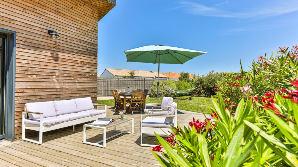 Imaginez une grande terrasse en bois, baignée de soleil. On y trouve un salon de jardin moderne aux lignes épurées, tout en blanc, parfait pour se détendre. Un peu plus loin, un coin repas s’installe à l’ombre fraîche d’un parasol vert vif. Et l’ensemble s’ouvre sur un jardin généreux : une belle pelouse, un hamac qui invite à la sieste, et des lauriers-roses en pleine floraison. Un vrai havre de paix.