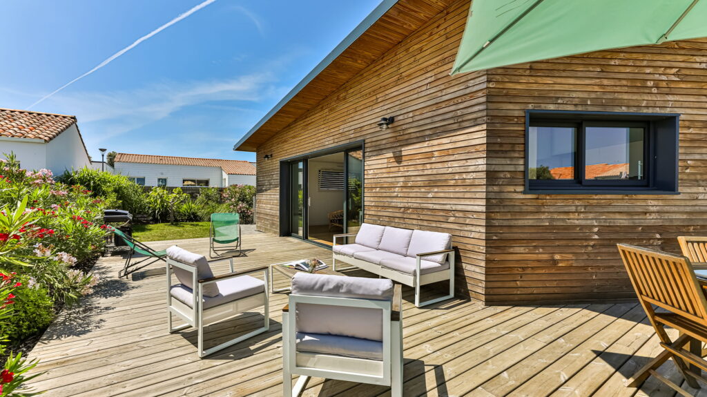 Imaginez une maison moderne, toute habillée de bois chaleureux. Devant, une grande terrasse invite à la détente, avec un coin salon confortable installé juste là, comme pour profiter pleinement de ce beau ciel bleu et ensoleillé.