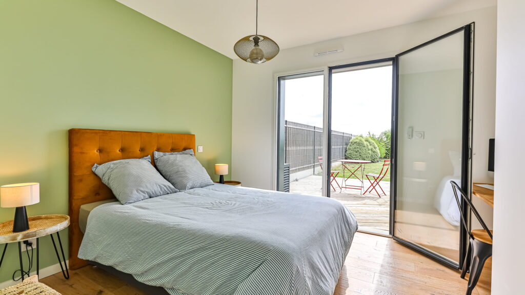 Chambre lumineuse avec un mur d'accent vert sauge, un lit double à tête de lit moutarde, et une grande baie vitrée ouverte sur une terrasse en bois.