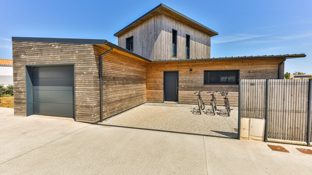 Cette maison moderne arbore un bardage en bois clair, qui contraste joliment avec la porte de garage gris anthracite. La porte d'entrée reprend la même teinte, créant une belle harmonie. Devant, trois vélos sont posés sur la cour en gravier, le tout baigné par la lumière d'un beau ciel bleu.