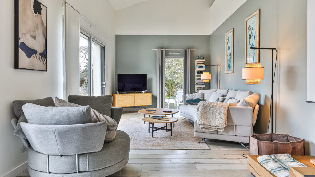 Imaginez un salon baigné de lumière, où le soleil caresse doucement un grand fauteuil rond gris, invitant à la détente. Un canapé aux tons clairs, enveloppé d'un plaid douillet, fait face à deux tables basses gigognes en bois qui apportent une touche chaleureuse. La télévision, discrètement posée sur un meuble en cannage, ne perturbe pas la sérénité des lieux. De grandes baies vitrées ouvrent l'espace et laissent entrer à flots la douce lumière du jour.