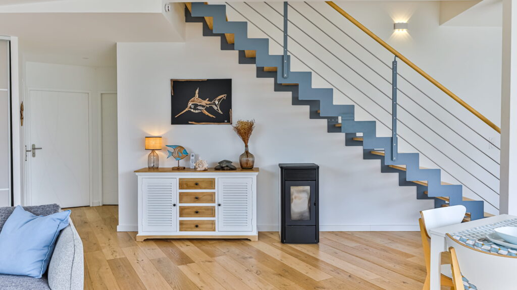Intérieur moderne lumineux avec un escalier ouvert en métal gris et bois. Sous l'escalier se trouvent un buffet blanc et bois avec une décoration d'inspiration marine, un tableau de requin, et un poêle à granulés noir.