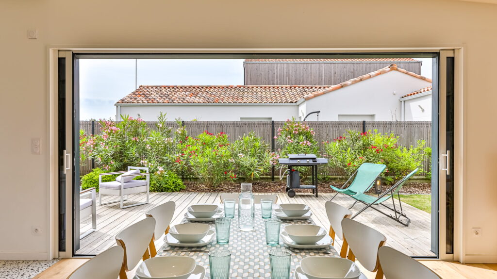 Table de salle à manger dressée face à une grande baie vitrée grande ouverte, offrant une vue dégagée sur une terrasse en bois avec barbecue, chaises longues et un jardin fleuri.