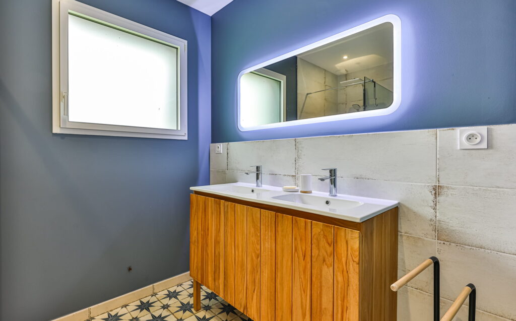 Meuble double vasque en bois clair surmonté d'un grand miroir LED rétroéclairé, dans une salle de bain aux murs bleu ardoise et au sol en carreaux de ciment à motifs.