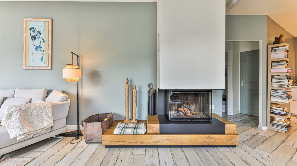 Voici un salon moderne et lumineux, où la cheminée contemporaine à foyer fermé, posée sur un socle en bois, attire immédiatement le regard. Sur la gauche, un canapé clair et un lampadaire élégant invitent à la détente. À droite, une haute étagère déborde de livres, ajoutant une touche chaleureuse et personnelle à l'ensemble.