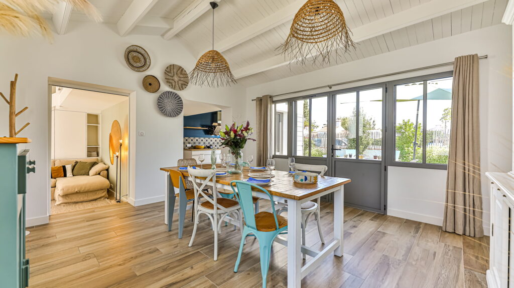 une salle à manger baignée de lumière, où l'esprit bohème chic prend vie. Une grande table en bois massif, patinée par le temps, trône au centre, entourée de chaises dépareillées qui racontent chacune leur histoire. Au-dessus, le plafond en lambris blanc met en valeur de belles poutres apparentes, ajoutant une touche de caractère. Et pour couronner le tout, de grandes portes-fenêtres grises s'ouvrent sur un jardin extérieur, comme une invitation permanente à la détente.