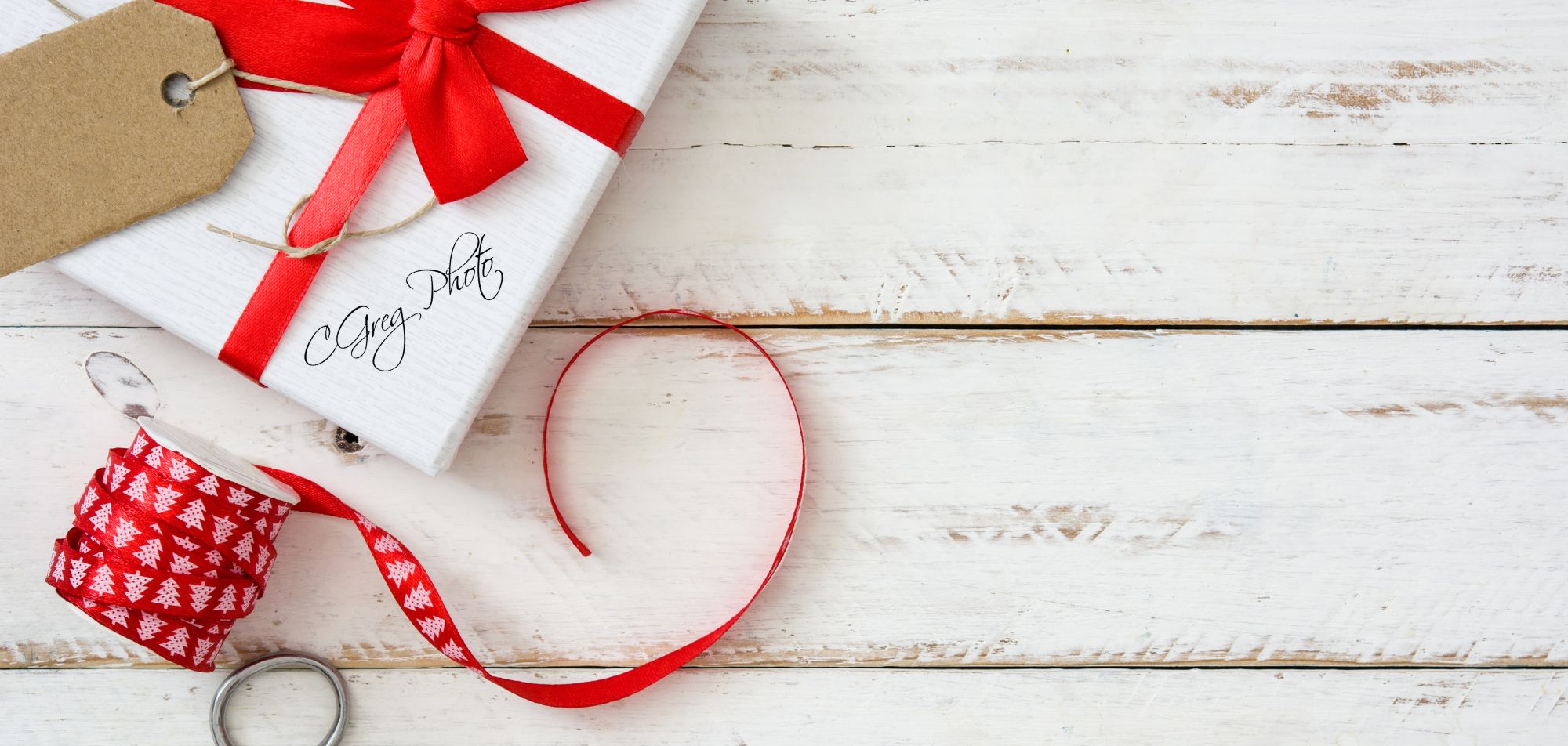 carte cadeau 1 Sur une vieille table en bois blanc, un paquet cadeau blanc avec un joli nœud rouge attend. À côté, une étiquette en kraft toute simple, et un rouleau de ruban de Noël rouge décoré de petits sapins. La table est dégagée sur la droite, comme si on avait laissé de la place pour poser autre chose.