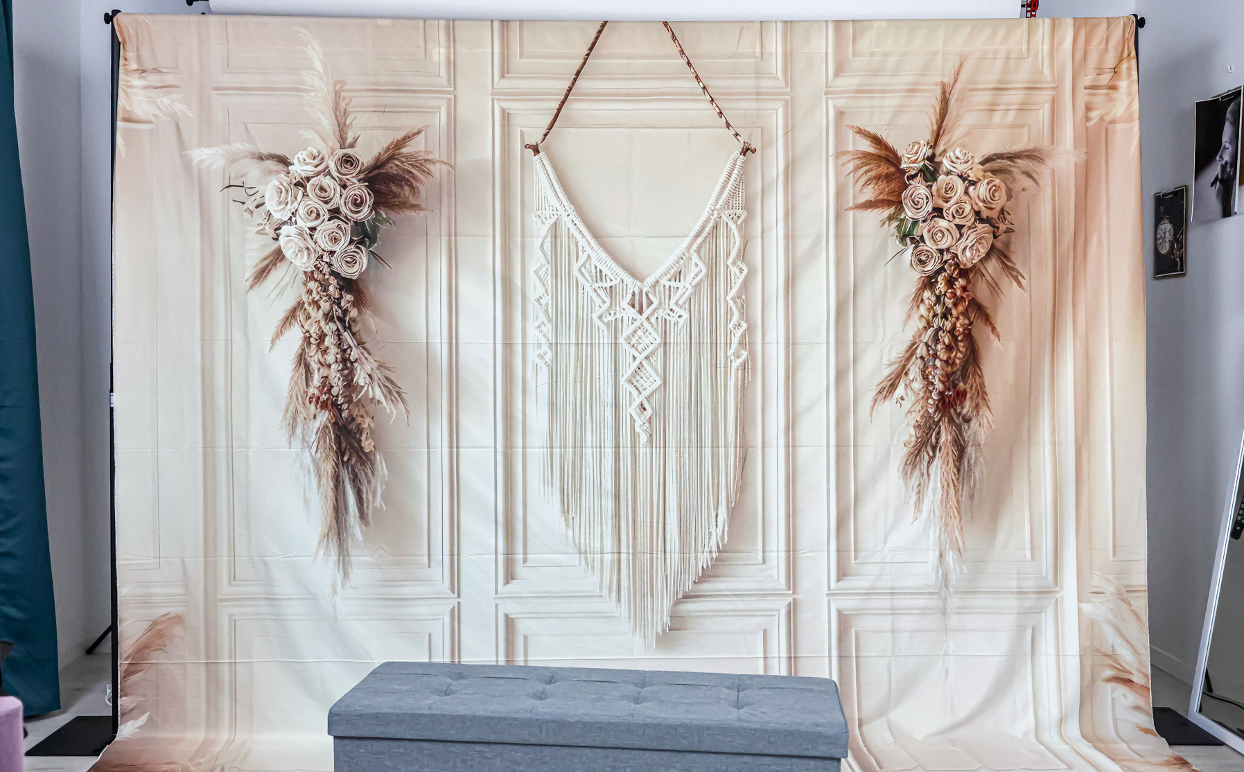 Dans ce décor de studio photo au style bohème, on trouve une toile de fond imprimée qui imite des moulures classiques. Au milieu, une grande tenture en macramé attire le regard, entourée de chaque côté par des bouquets symétriques de fleurs séchées, avec des roses et de l'herbe de la pampa. Devant, un petit banc capitonné gris est placé au premier plan, prêt à être utilisé.