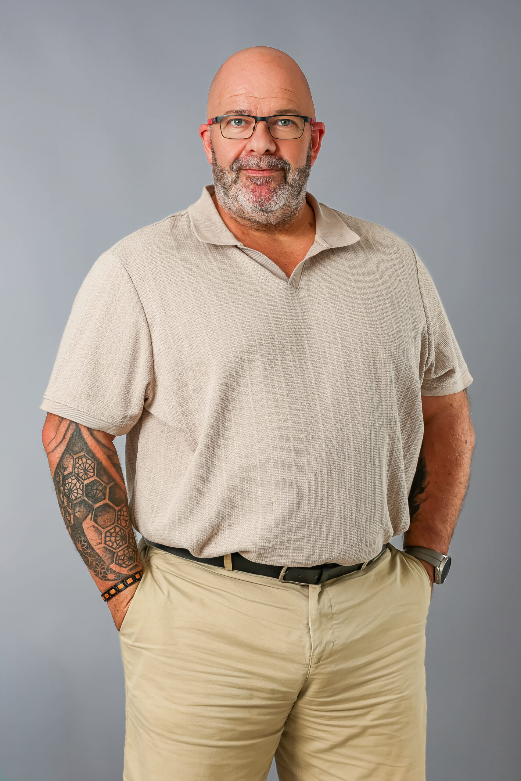 Portrait en studio à mi-corps d’un homme d’âge mûr, chauve et barbu, portant des lunettes, qui regarde l'objectif avec un sourire serein. Il porte un polo beige texturé rentré dans un pantalon kaki avec une ceinture foncée, et a ses mains dans ses poches. Un grand tatouage géométrique de type alvéoles couvre son avant-bras droit, et il porte une montre de sport au poignet gauche. Le fond est un studio gris uni.